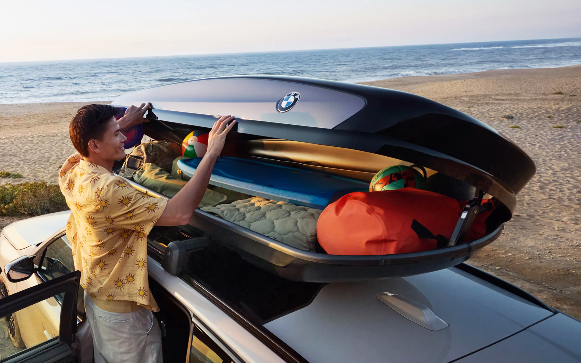 A man opens a BMW roof box filled with beach gear, with the ocean in the background.