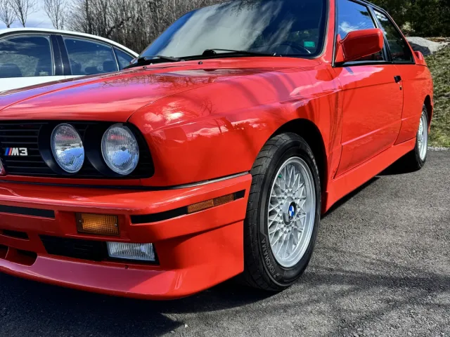 BMW M3 E30 rouge, trois quarts avant, jantes BBS visibles.