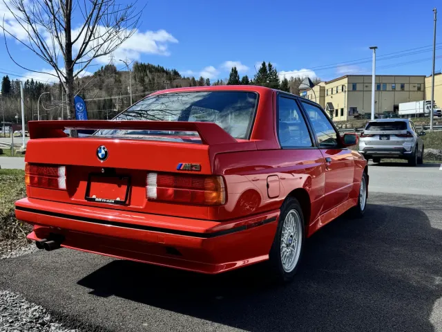 BMW M3 E30 rouge, trois quarts arrière, aileron M3 visible.