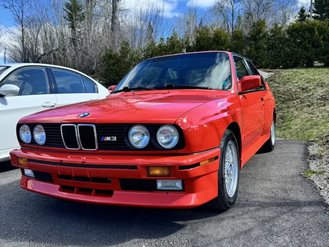 BMW M3 E30 rouge, trois quarts avant gauche, badge M3 sur l’aile.